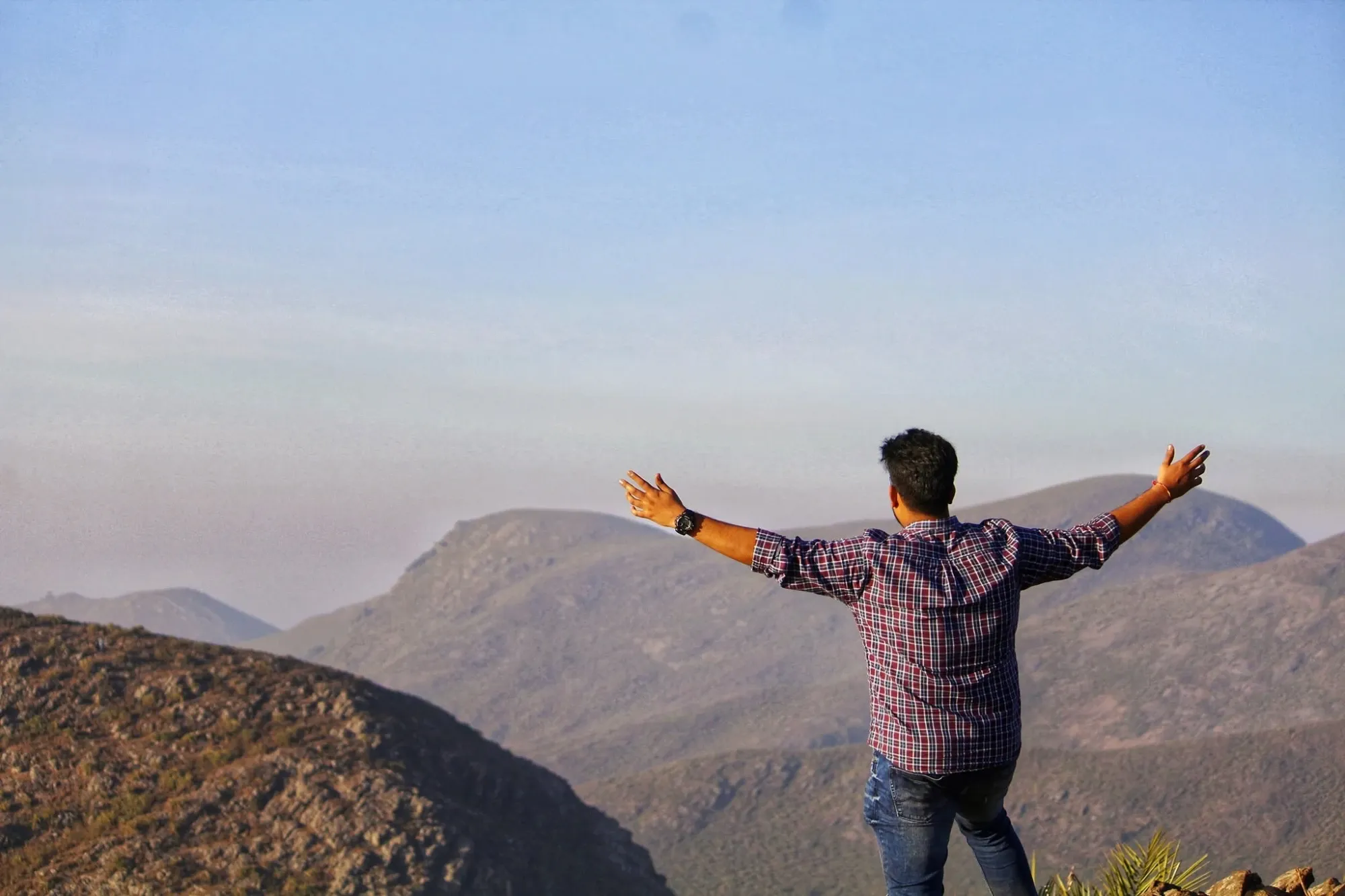 Rahul from behind with arms spread wide on a rocky hilltop above hazy hills